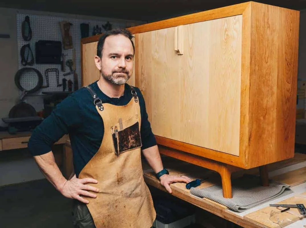 Michael McQuaid in his apron, standing beside a cherry cabinet in his studio