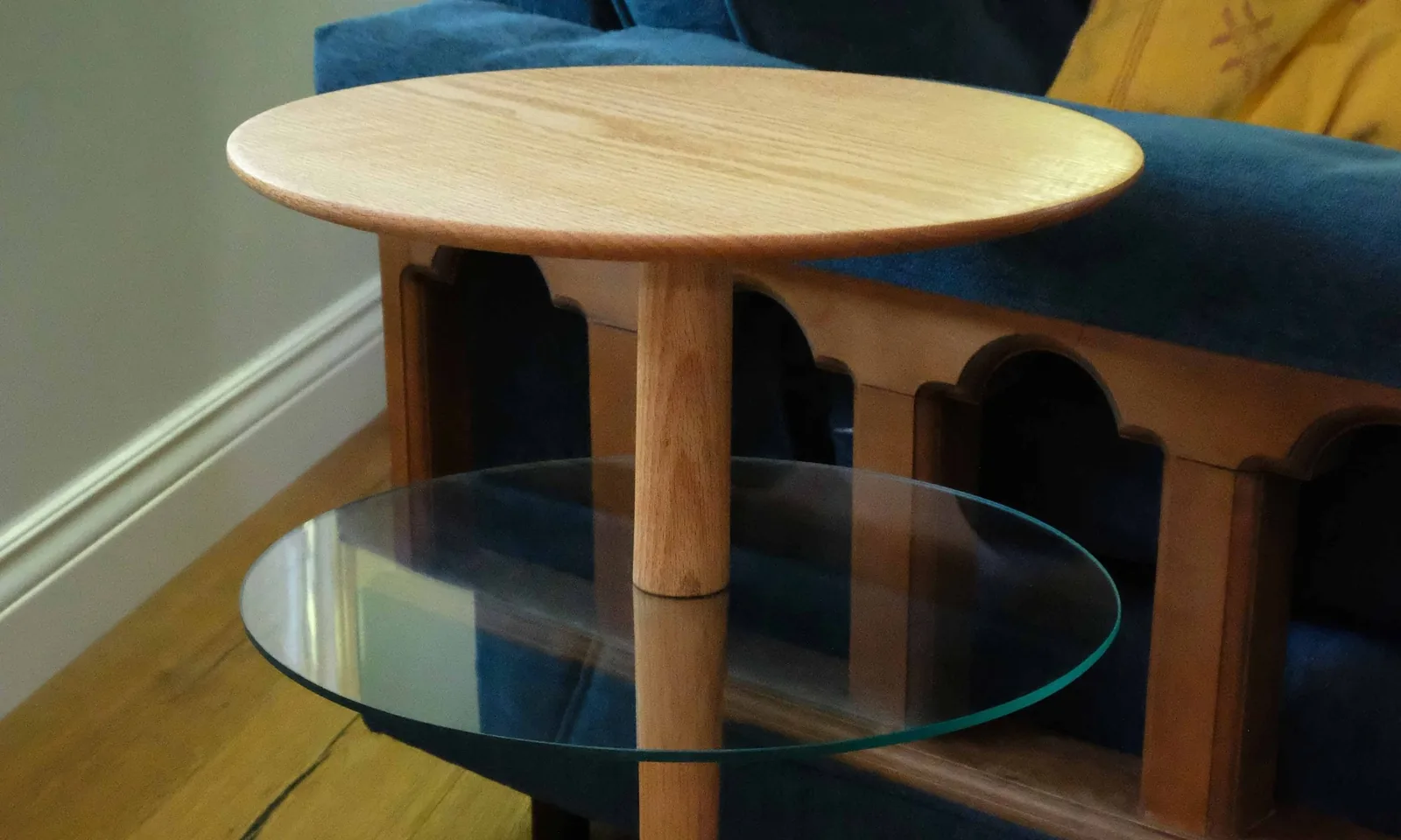 Round pedestal table with a glass lower shelf beside a blue sofa